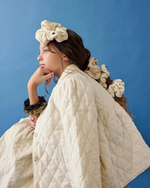 Profile view of a young girl with curly brunette hair wearing a scrunchie headband and multiple scrunchies. She is wearing a quilted jacket.