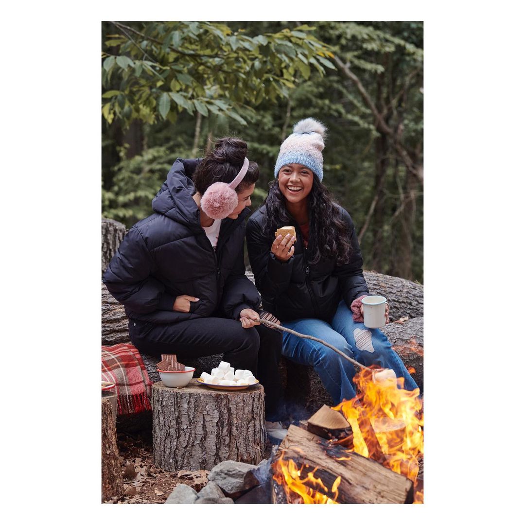 Two smiling women in winter clothing enjoy roasting marshmallows over a campfire in a wooded area, creating a cozy, outdoor scene.