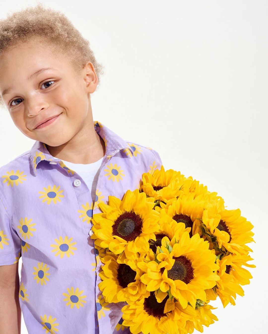 A happy child with curly blonde hair smiles while holding a bunch of yellow sunflowers against a clean white background.