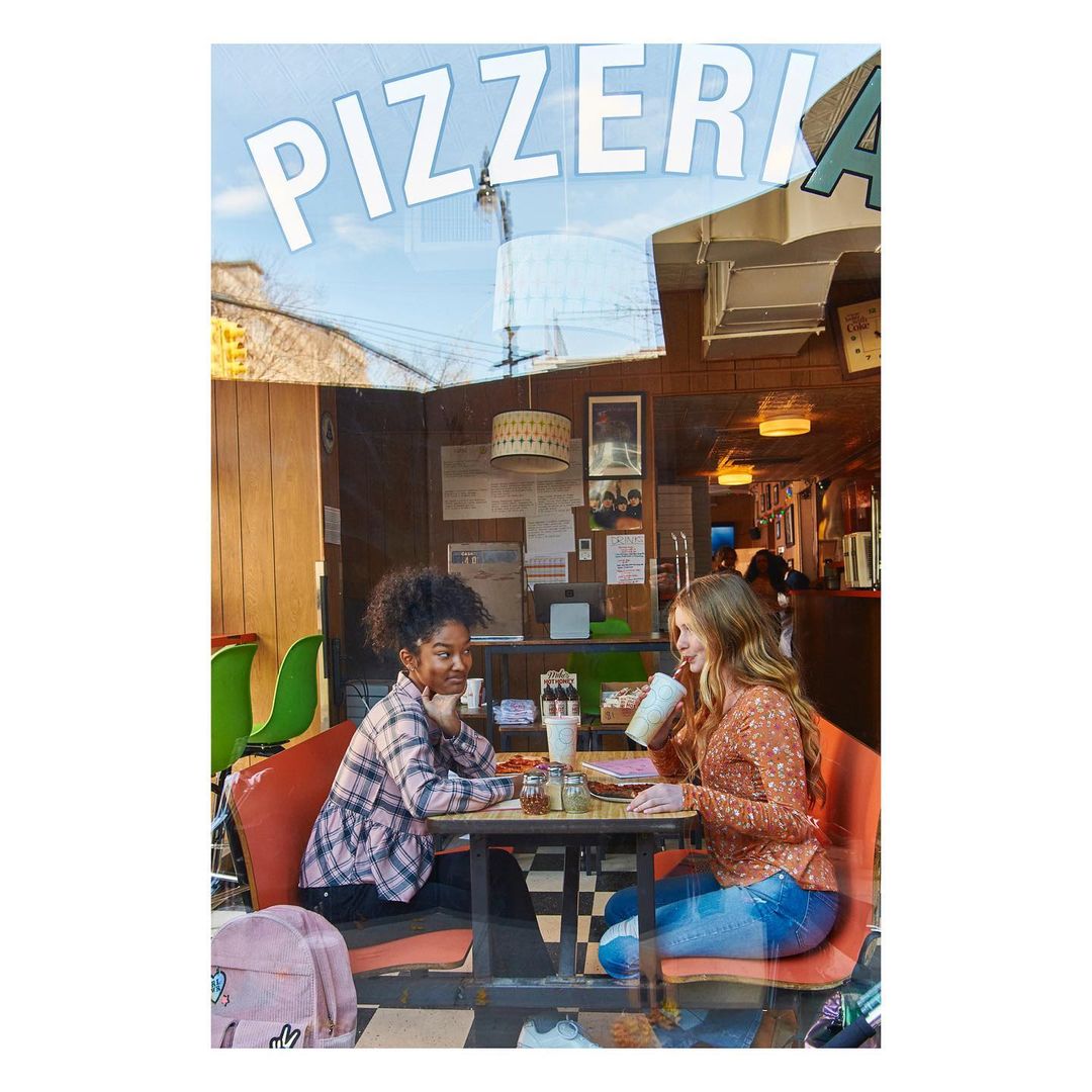 Two young girls seated at a table inside a pizzeria, sharing pizza and drinks. The pizzeria sign is visible in the window.