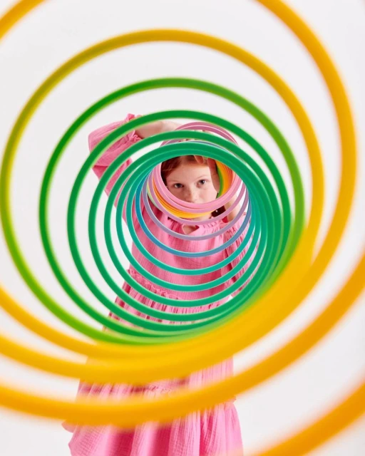 A child with red hair looks through a colorful slinky toy against a white background in a studio setting, creating a playful image.