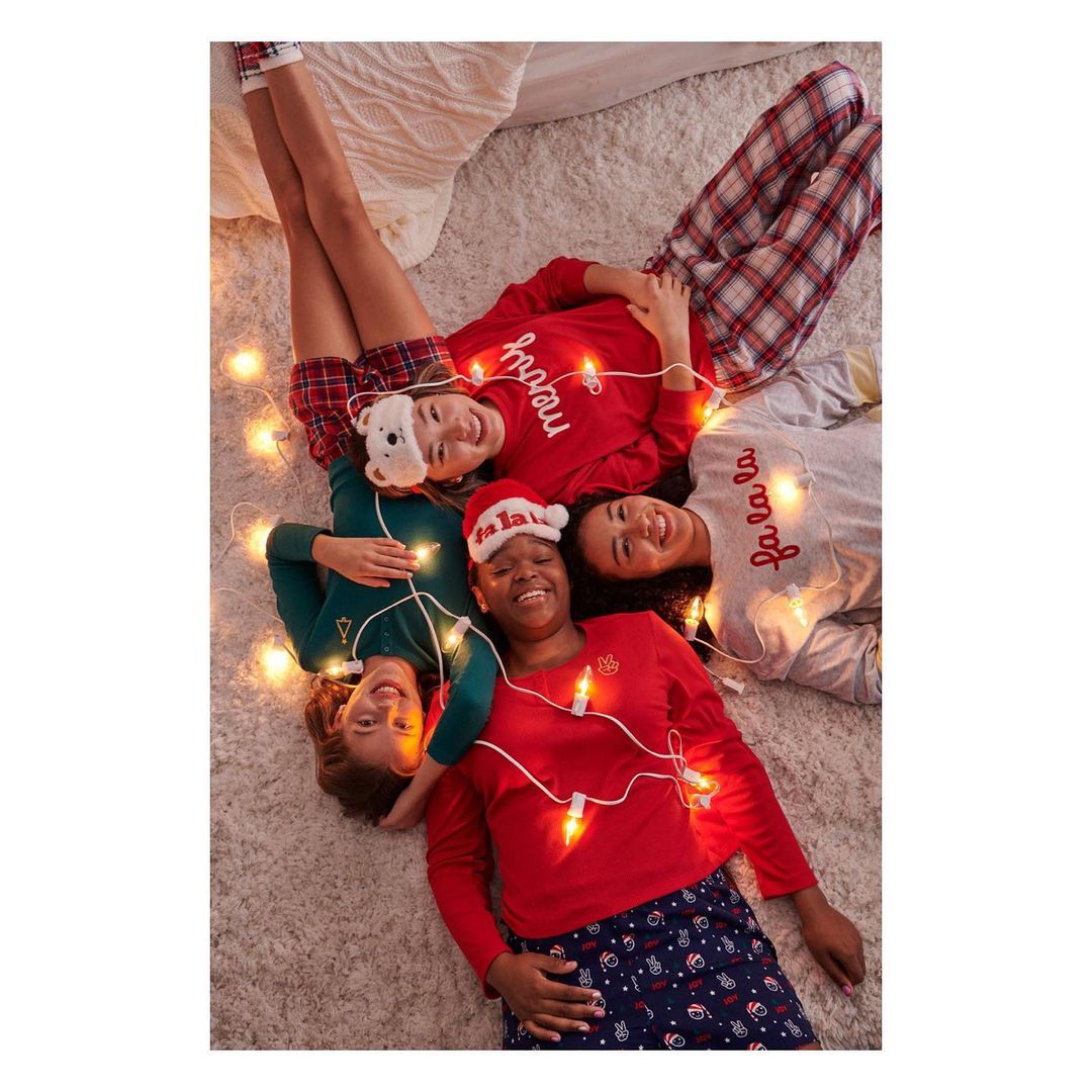Four smiling young women in pajamas lie on a rug surrounded by string lights; a group enjoying a cozy, festive indoor gathering.