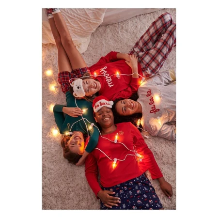 Four smiling young women in pajamas lie on a rug surrounded by string lights; a group enjoying a cozy, festive indoor gathering.