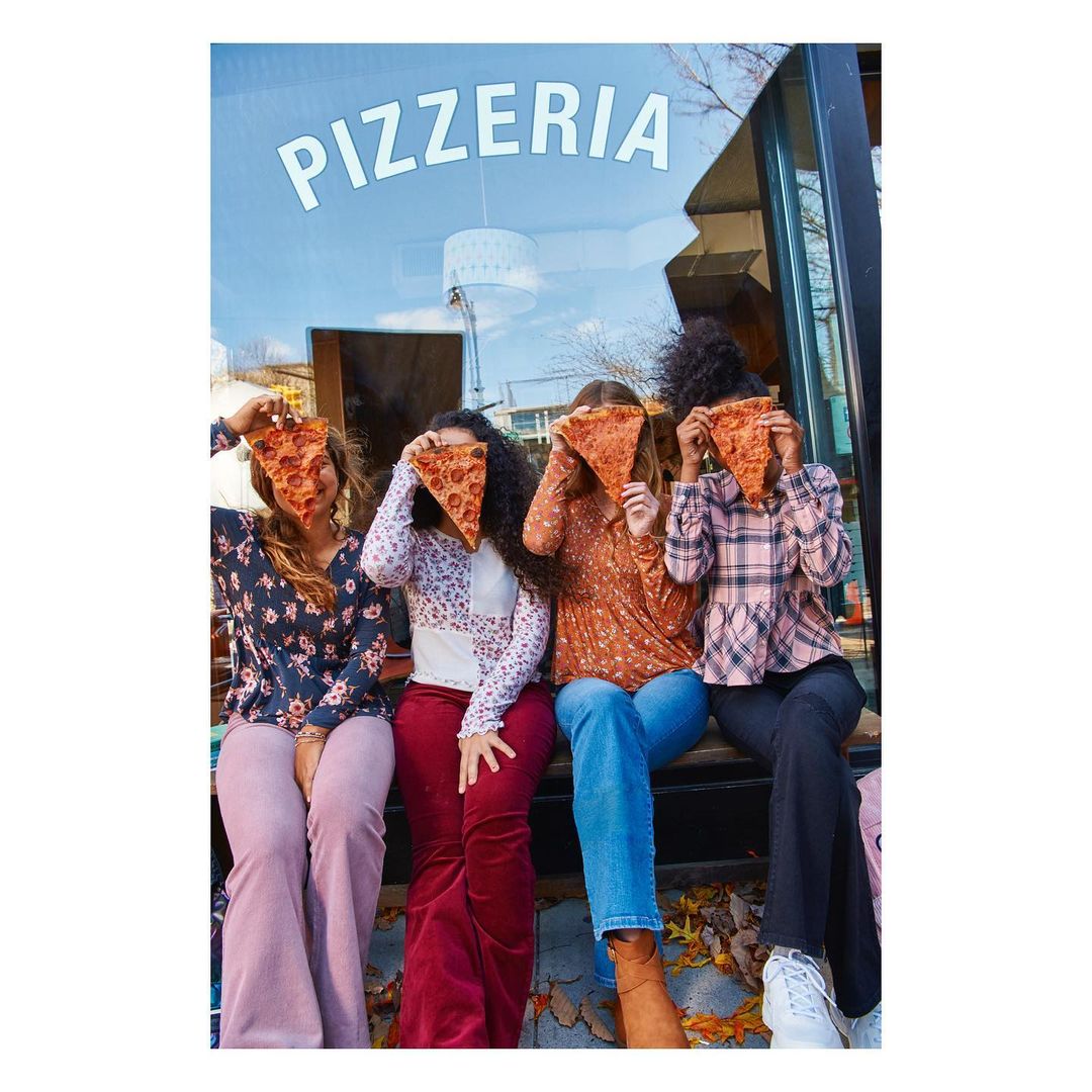 Four young women seated outdoors in front of a pizzeria, holding slices of pizza in front of their faces in a fun, casual pose.
