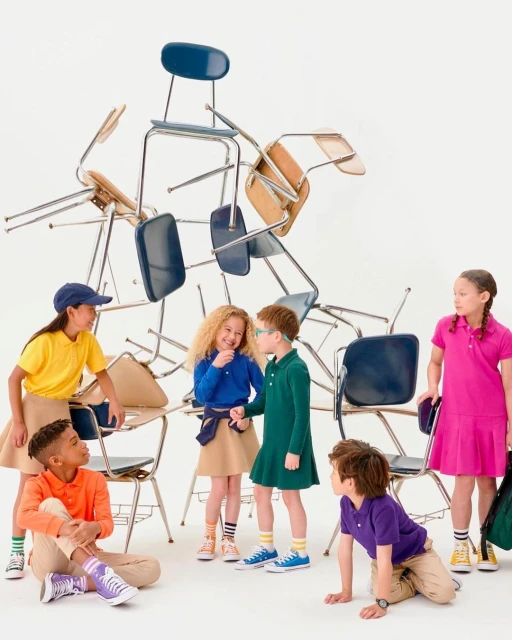 Group of six children wearing colorful polo shirts and khaki pants/skirts posing near a pile of blue and tan school chairs on a white background.