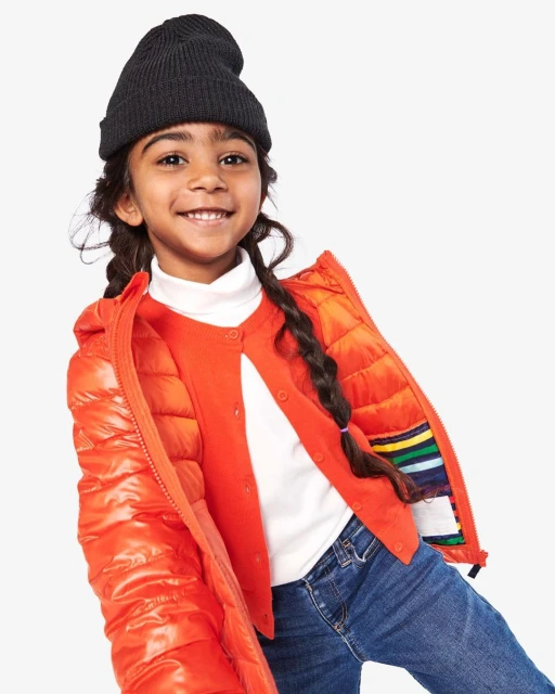 A young girl smiles brightly in a studio shot, wearing a black beanie, orange puffer jacket, red sweater, and jeans against a white backdrop.