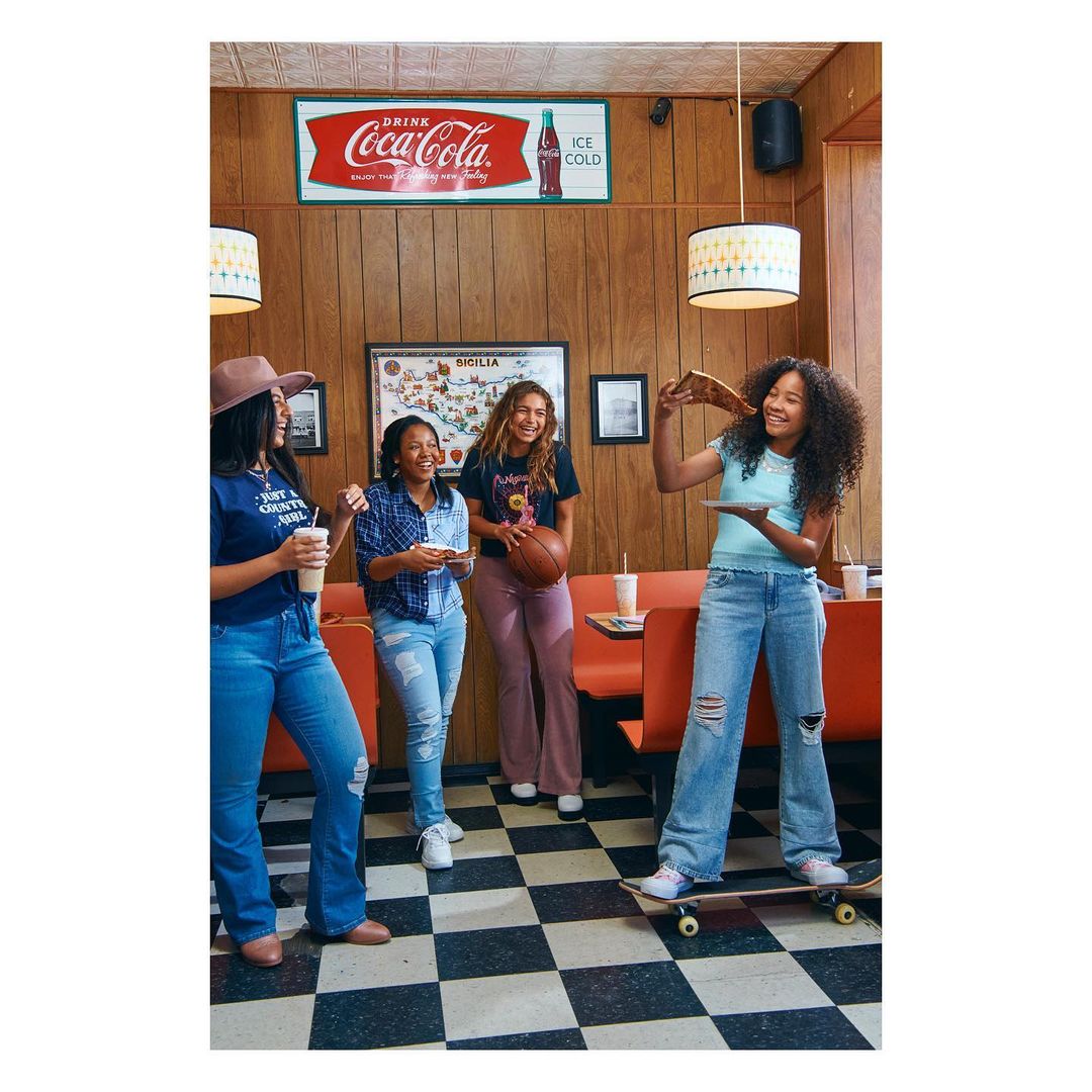 Four young women in casual clothes at a diner. One on a skateboard holding pizza, retro decor, Coca-Cola sign, checkerboard floor.