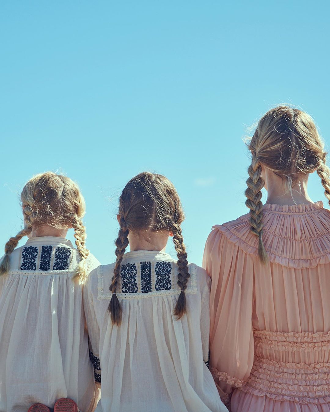 Three young girls with blonde hair styled in braids stand close together in matching dresses against a bright blue sky backdrop.
