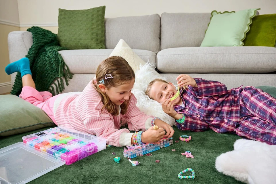 Two young girls in pajamas lying on a green rug indoors, making bracelets from colorful rubber bands and plastic looms.