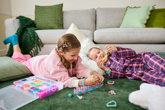 Two young girls in pajamas lying on a green rug indoors, making bracelets from colorful rubber bands and plastic looms.