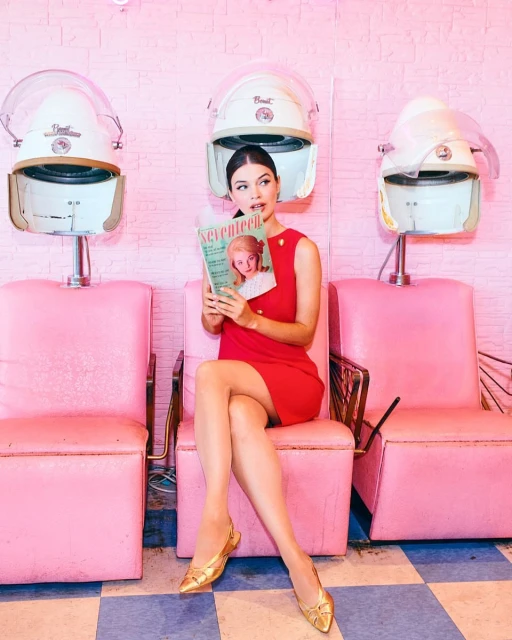 Glamorous woman in a red dress sits in a pink salon chair, reading a vintage Seventeen magazine under a hair dryer, vintage retro aesthetic.
