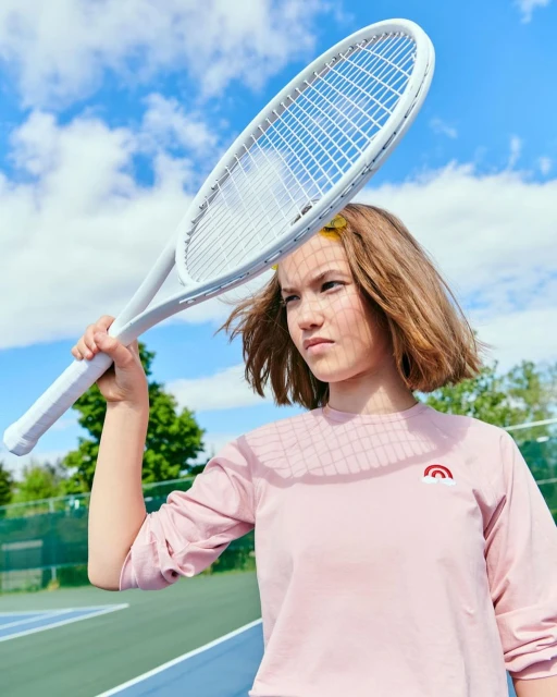 Teen girl poses on tennis court holding up a white tennis racket against a blue sky with clouds. She wears a pink shirt.