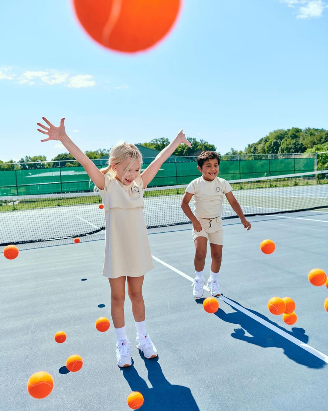 A young girl with arms raised, accompanied by a boy, is playing on a tennis court with orange tennis balls against a blue sky backdrop.