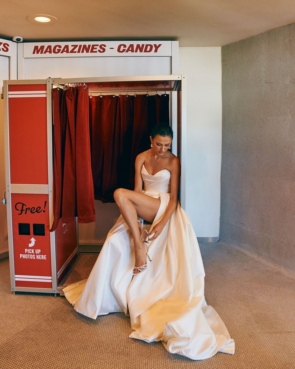 Woman in a strapless white gown and high heels poses in front of a red and white photo booth. She is sitting and putting on her shoe.