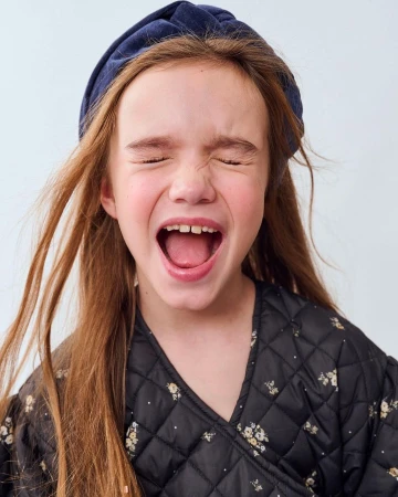 Close-up of a smiling young girl with long red hair wearing a navy blue headband and floral jacket against a white background.
