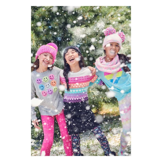 Three smiling young girls stand outdoors surrounded by falling snow. They are wearing colourful winter hats and sweaters.