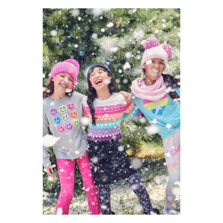 Three smiling young girls stand outdoors surrounded by falling snow. They are wearing colourful winter hats and sweaters.