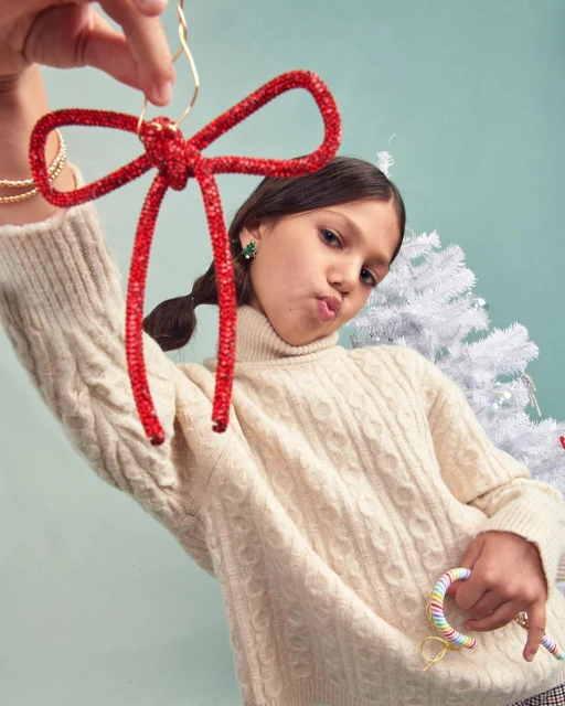 Young girl in cable-knit sweater poses with a red decorative bow and rainbow keychain against a blue backdrop with Christmas tree.