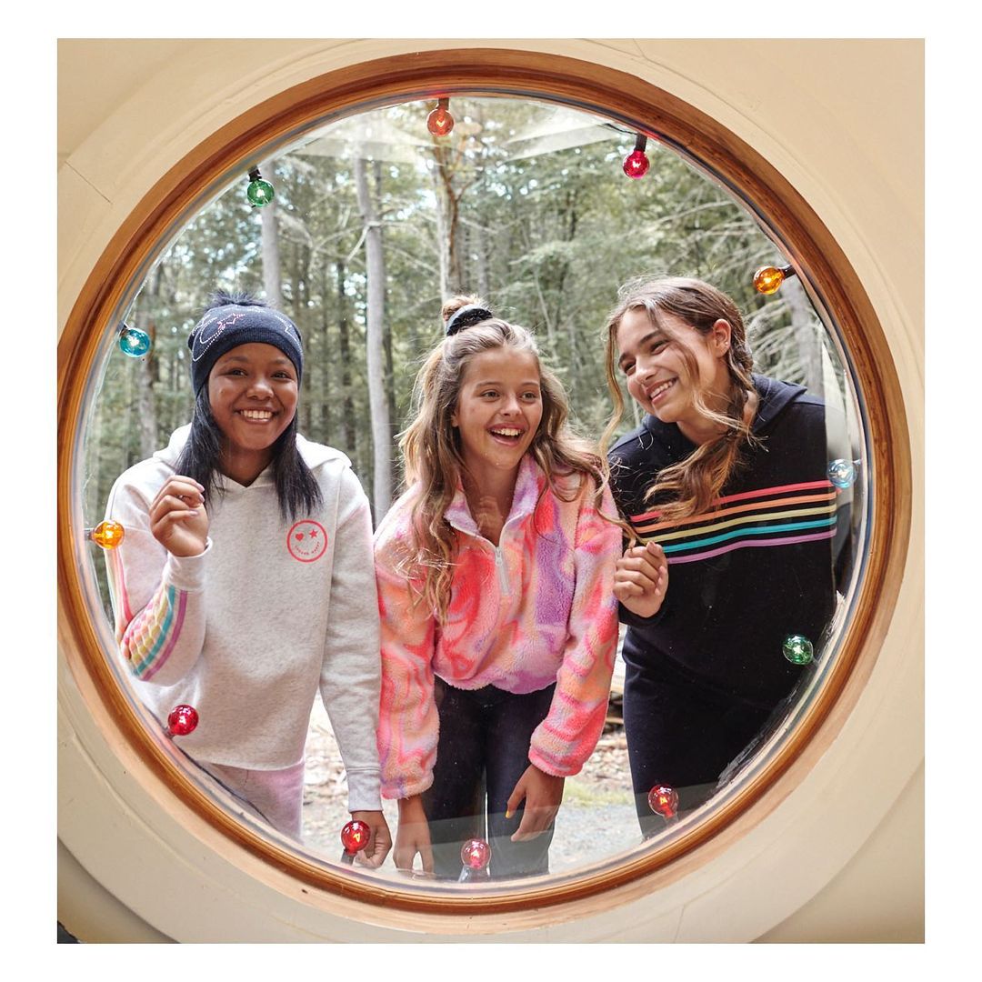 Three smiling girls in hoodies pose inside a whimsical round treehouse window overlooking a forest, adorned with colorful decorative lights.
