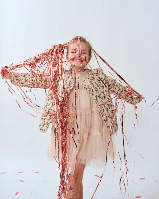 Young girl smiling and playing with metallic confetti streamers in a studio setting. She wears a floral jacket and peach-colored dress.