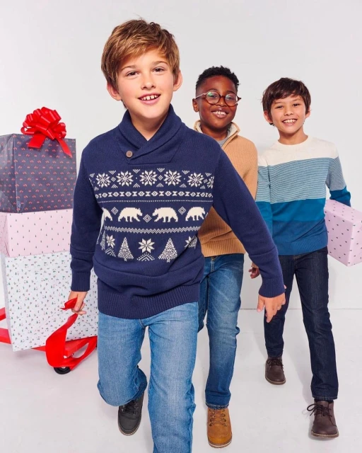 Three boys smiling and walking indoors, wearing patterned sweaters and jeans, carrying wrapped gifts against a white background.