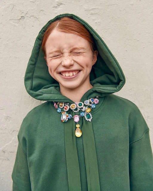 Red-haired child in a green hooded sweatshirt smiles with her eyes closed, wearing a colorful jeweled necklace against a neutral wall.