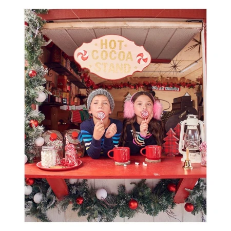 Two children at a holiday hot cocoa stand, wearing winter clothing and holding spiral lollipops, with Christmas decorations surrounding them.