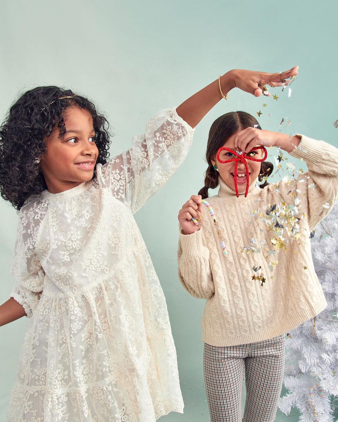 Two young girls pose indoors with confetti, festive holiday glasses, and a white Christmas tree. One wears a lace dress, the other a sweater and patterned pants.