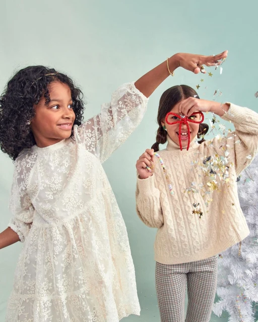 Two young girls pose indoors with confetti, festive holiday glasses, and a white Christmas tree. One wears a lace dress, the other a sweater and patterned pants.