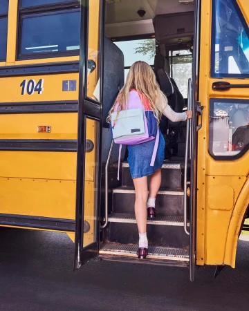A young student with a backpack is seen from behind, climbing the stairs of a classic yellow school bus on a sunny day.