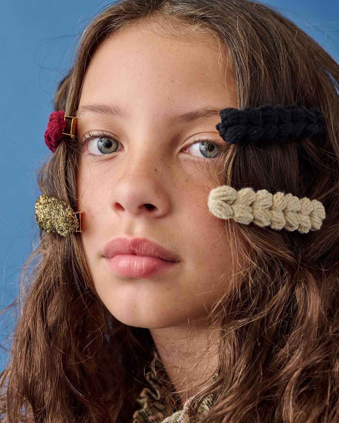 Close-up portrait of a young girl with brown wavy hair and colorful braided hair clips, against a solid blue background.