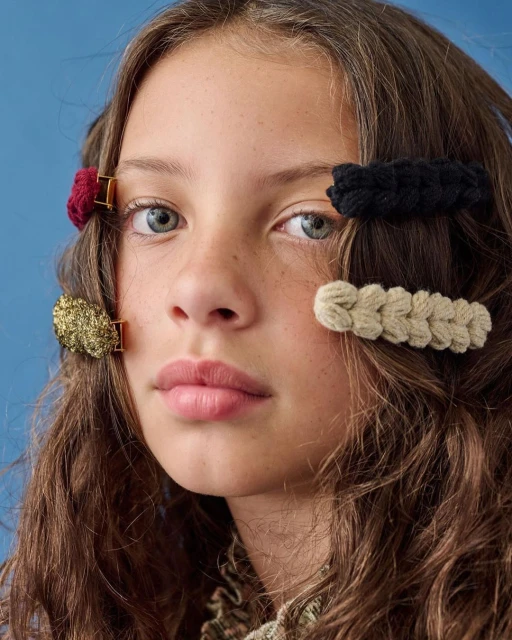 Close-up portrait of a young girl with brown wavy hair and colorful braided hair clips, against a solid blue background.