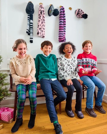 Four smiling children sit on a bench in front of a white wall with hats and scarves hanging, creating a festive holiday scene.