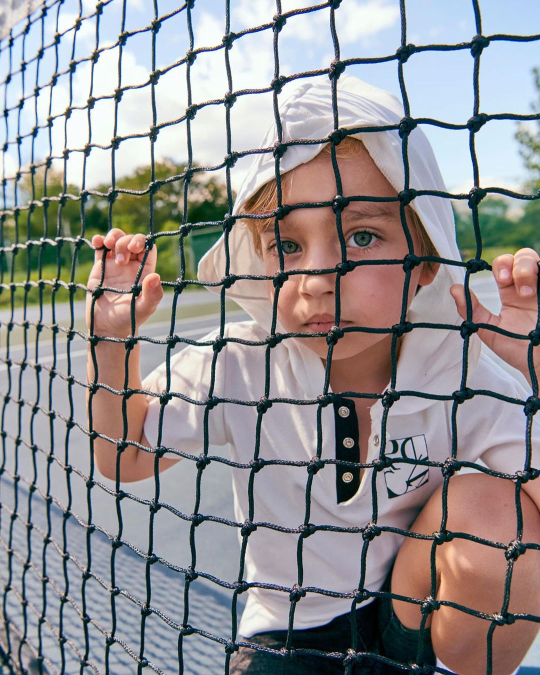 Young child in a hooded shirt peers through a black net on a tennis court with trees and a light blue sky in the background.