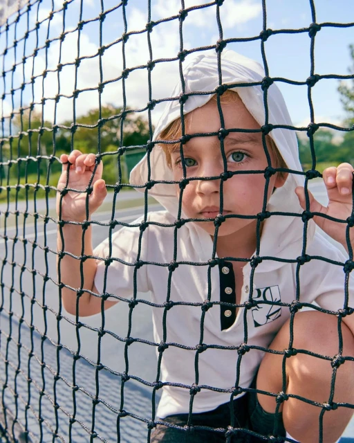 Young child in a hooded shirt peers through a black net on a tennis court with trees and a light blue sky in the background.