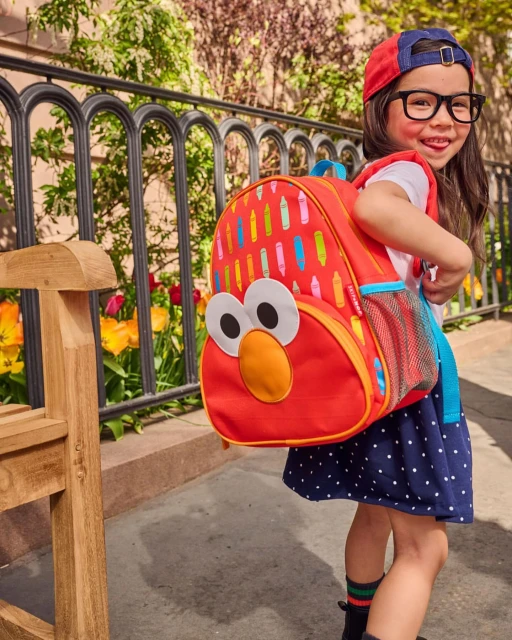 Smiling child with Elmo backpack outdoors on sunny day, wearing glasses and a cap, next to a bench and flowers in the background.