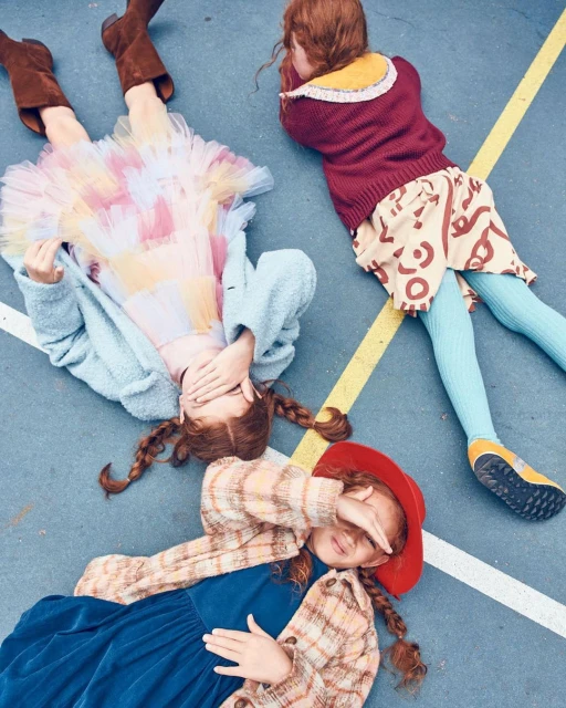 Three young girls with red hair are lying on a blue surface, with two of them covering their faces with their hands.