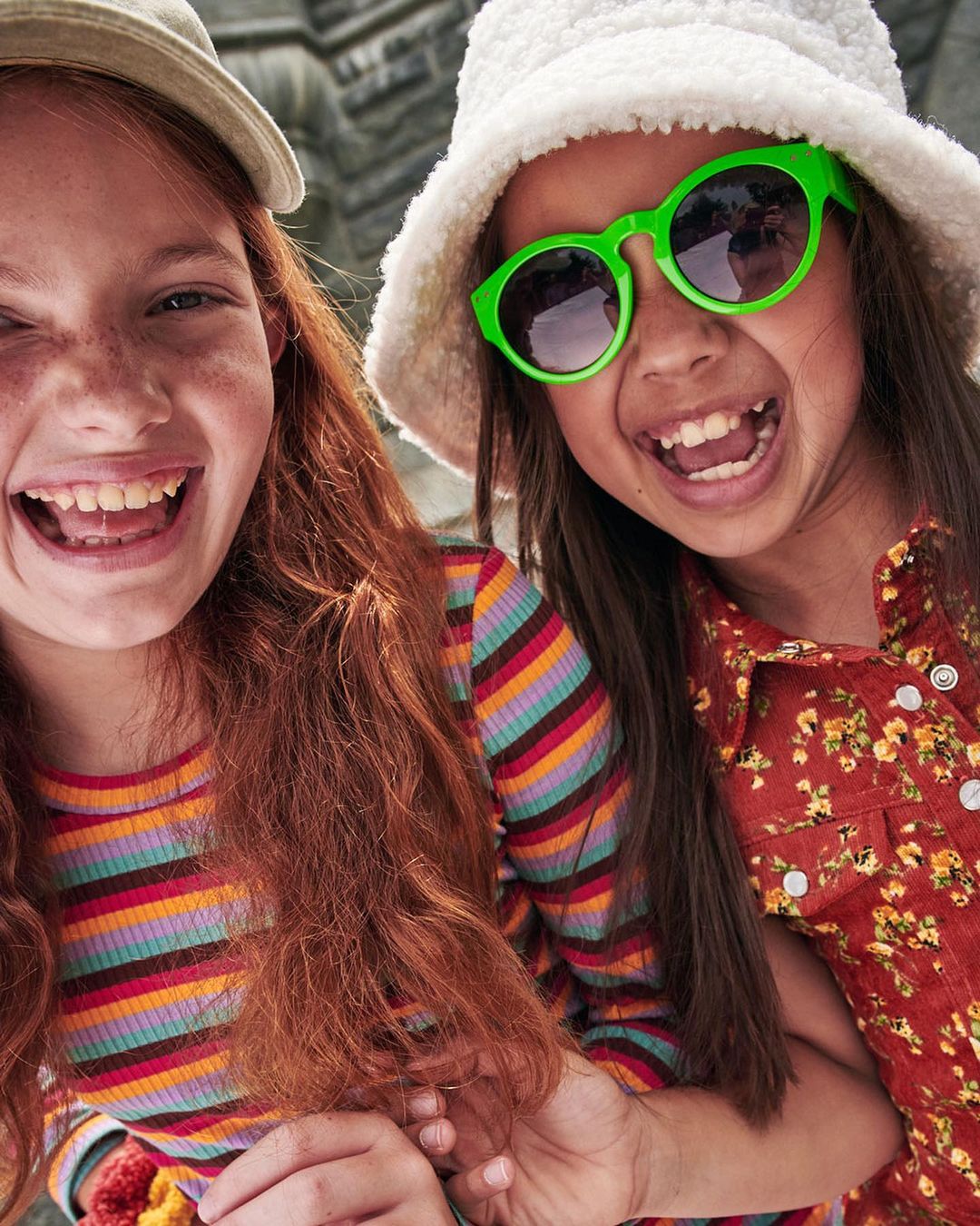 Two smiling girls in bright clothes and hats pose closely together, showcasing childhood fashion against a blurred background.