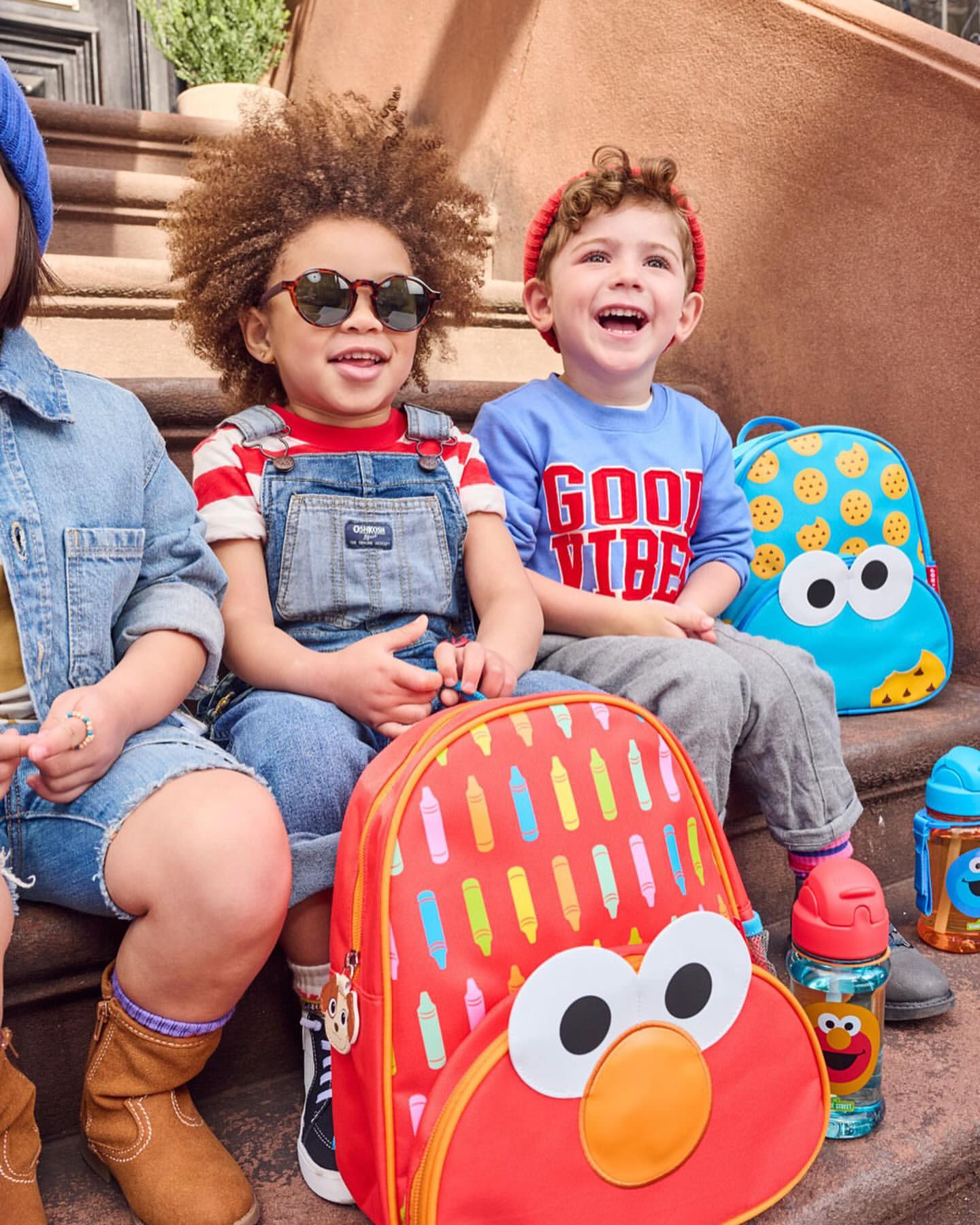 Three young children sit on outdoor steps with Sesame Street backpacks. The kids wear casual outfits, and natural light illuminates the scene.