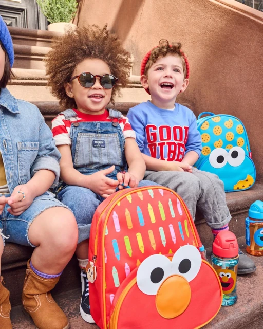 Three young children sit on outdoor steps with Sesame Street backpacks. The kids wear casual outfits, and natural light illuminates the scene.
