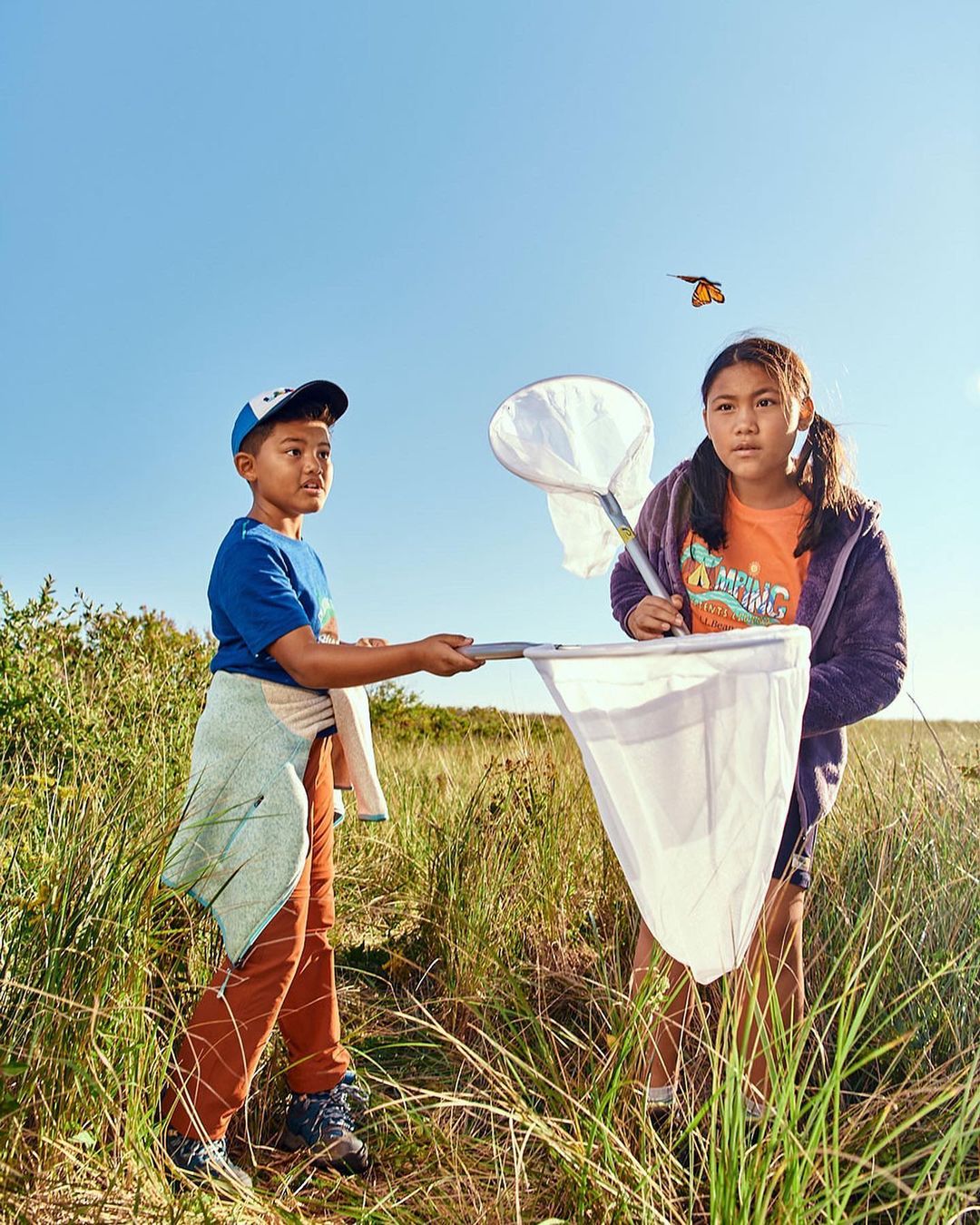Two Asian children stand outdoors in tall grass with a butterfly net trying to catch an orange monarch butterfly against a clear blue sky.