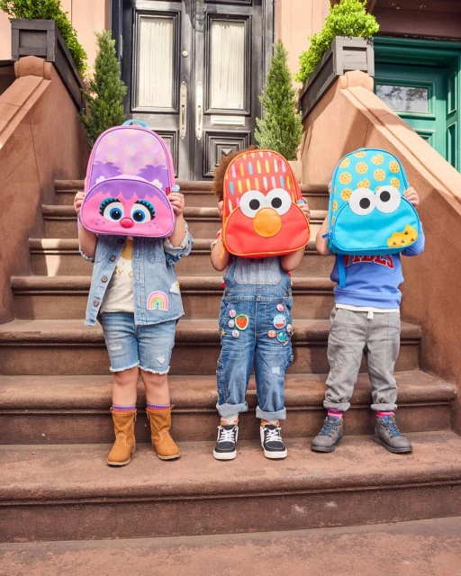 Three kids stand on stairs holding character backpacks, featuring Elmo, Cookie Monster, and Abby Cadabby from Sesame Street.