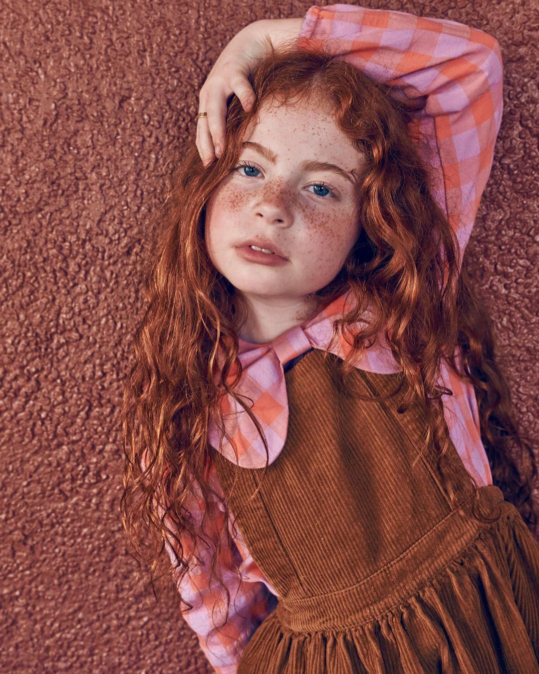Young redhead girl with freckles lies on a textured surface wearing a brown corduroy jumper and pink plaid top in a portrait shot.