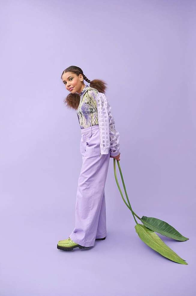 Young person in a printed shirt and lilac pants poses with large green leaves against a lilac background in a studio setting.