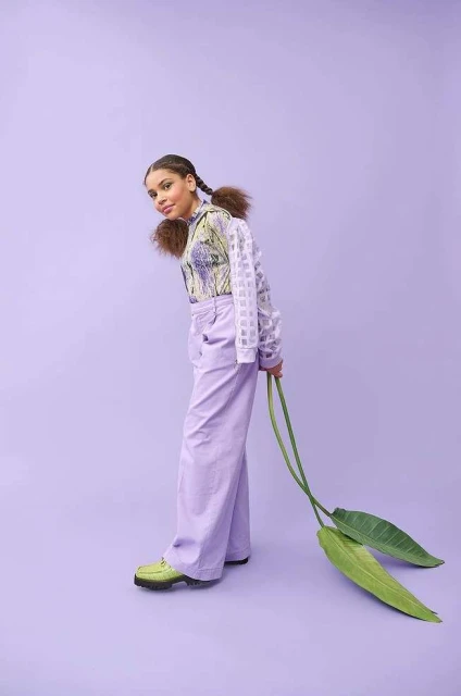 Young person in a printed shirt and lilac pants poses with large green leaves against a lilac background in a studio setting.