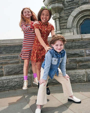 Three young girls posing playfully together in front of a weathered stone architectural structure outdoors.