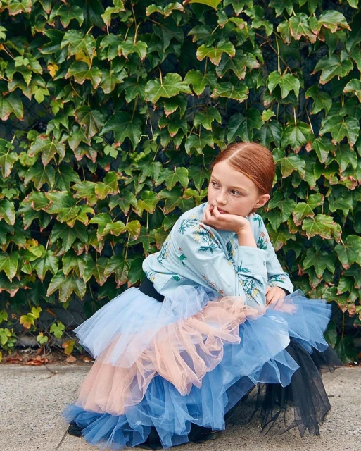 Young girl with red hair sits in front of an ivy-covered wall, wearing a light blue floral blouse and layered tutu in blue, peach, and black.