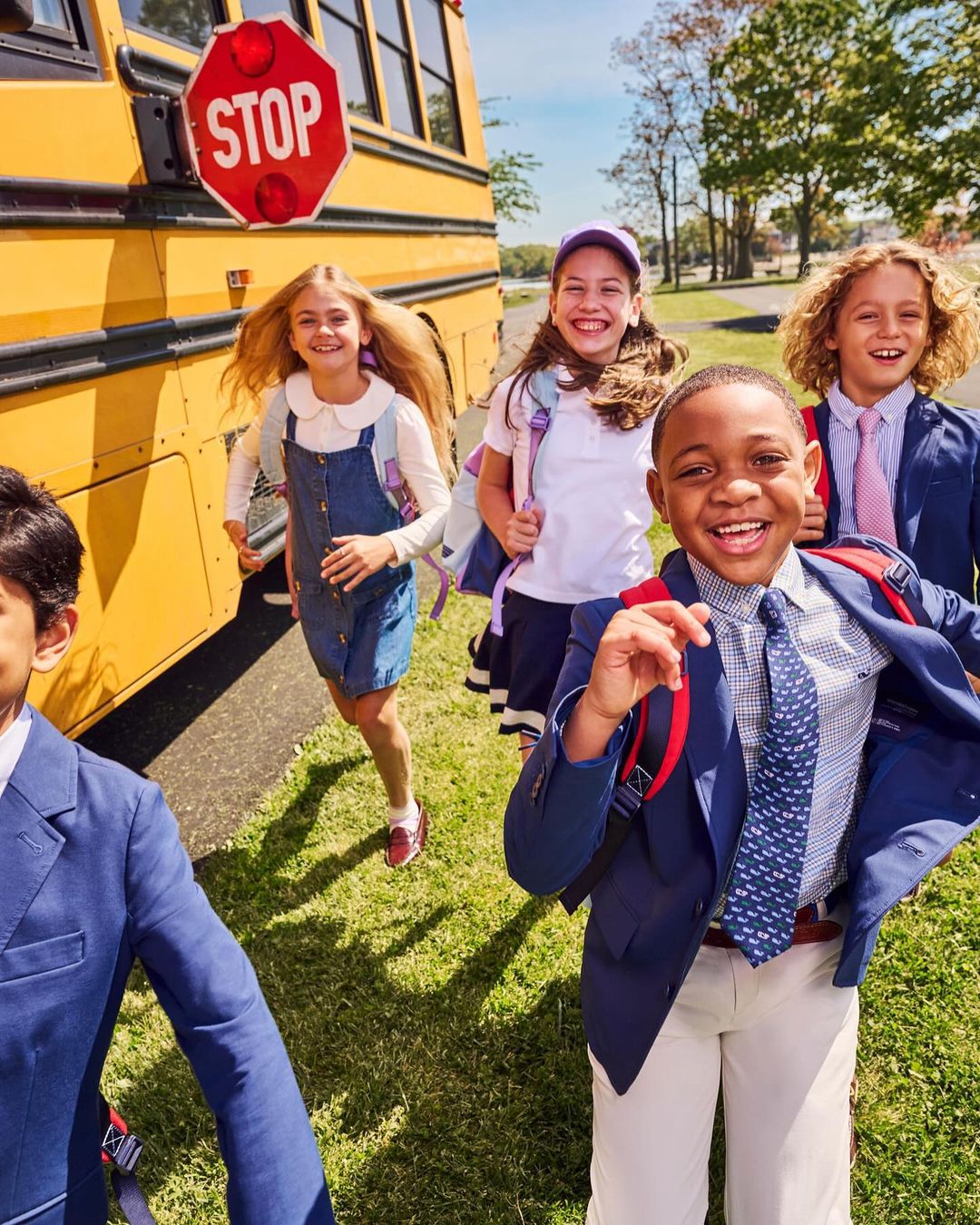 A diverse group of children exit a yellow school bus near a red stop sign, all wearing blue blazers and backpacks, smiling and running on green grass.