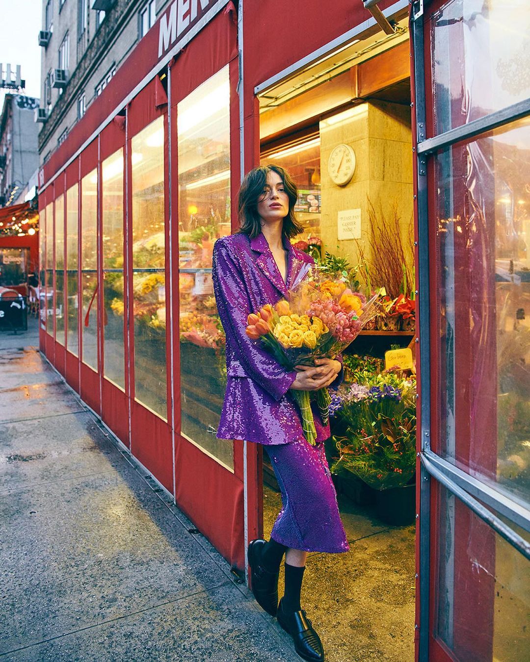 Fashionable woman in a purple sequin suit stands holding a bouquet of flowers in front of a flower shop in an urban setting.