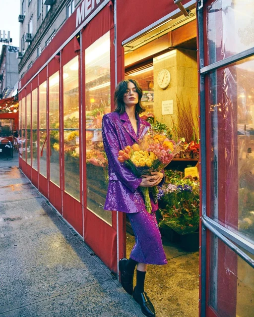 Fashionable woman in a purple sequin suit stands holding a bouquet of flowers in front of a flower shop in an urban setting.
